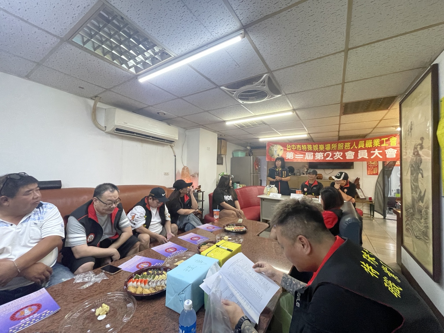 會員大會布條前，兩位與會者站立交流，其他與會者在旁聆聽